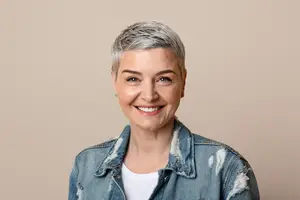 A woman with gray hair is wearing a denim jacket and a white shirt. She is smiling and seems to be posing for a photo.