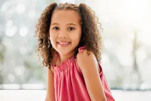 A young girl with curly hair wearing a pink dress is smiling and posing for a photo in a room with a bright, blurry background.