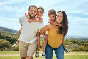 A man and a woman are carrying two kids in a field under a blue sky with mountains in the distance