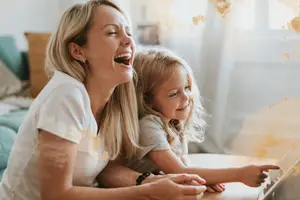 A woman and a young girl are laughing while lying on a bed and watching something on a tablet.