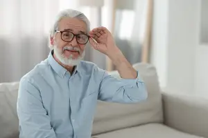 An elderly man with white hair and beard, wearing glasses and a blue shirt, sitting on a couch and adjusting his glasses with his right hand.
