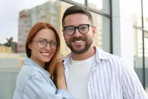 A couple with glasses smiling for a photo in front of a building