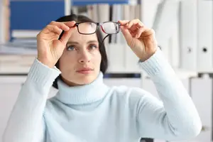 A woman wearing a blue turtleneck is adjusting her glasses.