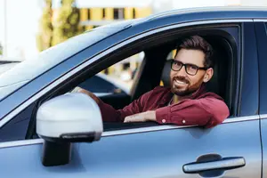 A man with a beard is smiling inside a blue car parked outside.