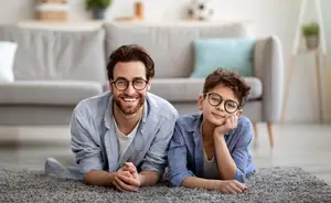 A man and a young boy sitting on the floor in a living room, both wearing glasses and smiling