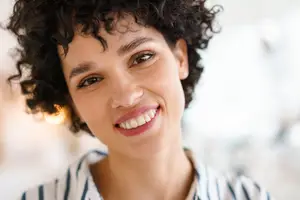 A woman with curly hair and a blue and white striped shirt is smiling at the camera with a bright light illuminating her face
