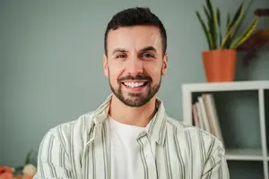 Smiling man with beard in a striped shirt posing in front of a green wall with plants
