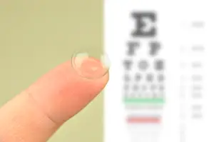 Close up of a finger holding a soft contact lens with a vision chart in the background