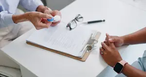 A man and a woman are sitting in front of a table with a document and glasses. The man is holding a blue and white object in his hand. The woman is holding the document. The man is wearing a ring, and the woman is wearing a watch. The table has a pen, a pencil, and a pair of glasses on it. The woman is wearing a ring, and the man is wearing a ring as well.