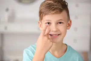 A boy wearing a blue shirt touches his eye with a finger.