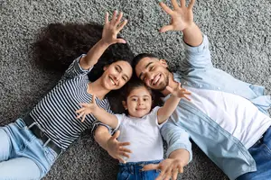 Three people lying on a carpeted floor with their hands raised, all smiling