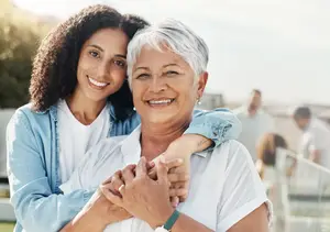 A smiling grandmother and granddaughter pose for a picture in an outdoor setting with other people in the background.