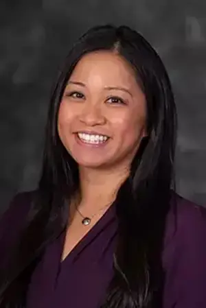 A smiling woman with long black hair wearing a purple blouse and a necklace