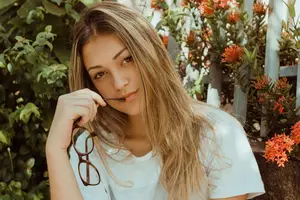 A young woman with blonde hair wearing a white shirt holding glasses