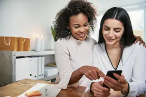 Two women are smiling and looking at a cell phone in an office room.