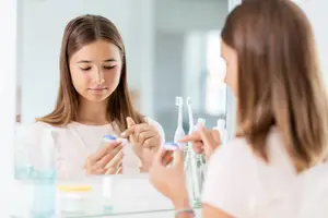 Two women looking in a mirror and applying toothpaste to their toothbrushes