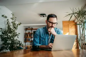 A man sitting at a desk with a laptop in front of him, wearing glasses and a blue denim shirt, with his right hand on his chin. He is looking at the laptop screen and seems to be working. Behind him is a shelf with books and a potted plant. On the right side is a wooden door with a glass window, and on the left side is a white wall with a potted plant on the floor.