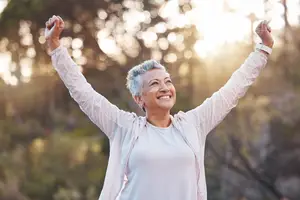 Happy senior woman standing in the forest with her arms raised