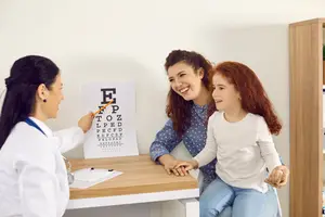 A doctor is performing an eye exam for a young girl with a smiling woman next to her.