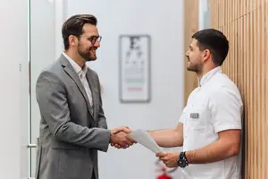Two men in business attire are shaking hands in an office hallway, one man is holding a piece of paper and wearing a watch.