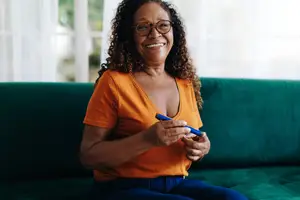 An older woman with curly hair wearing glasses and an orange shirt is smiling and holding a pen while sitting on a green couch.