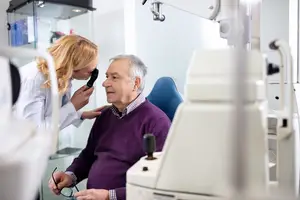 A female doctor in a white coat examining an elderly man's eyes using an ophthalmoscope in a clinic