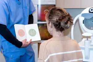 A woman at an eye clinic is looking at a book with circles while an optometrist holds a piece of paper