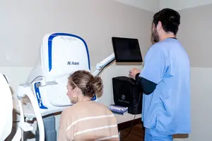 A woman is having her eyes examined by a man in a blue uniform in front of a Nikon device