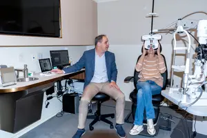A man and a woman are sitting in a room with medical equipment, the woman is wearing an eyepiece and the man is looking at a monitor