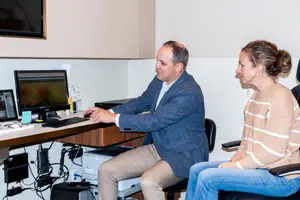 A man and woman sitting in chairs, the man is working on a computer, and the woman is looking at him