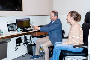 A man and a woman are sitting on chairs in an office room with monitors and a sink