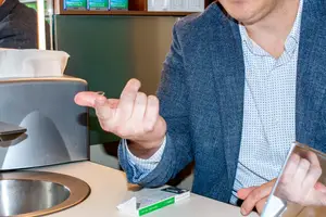 A man wearing a blue blazer is pointing at something with his finger while holding a tissue paper and a small object, possibly a ring, in his other hand. He is sitting in front of a sink with a stainless steel faucet and a tissue dispenser. On the right side, another man is holding a small object, possibly a phone, in his hand. Behind them, there is a wooden cabinet with several items on top.