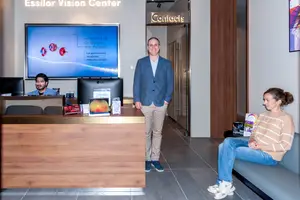 Three people in an eye care facility, with one man at the reception desk, another man behind the desk, and a woman sitting on a bench.