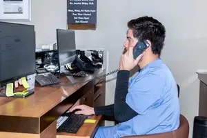 A man wearing a blue uniform is sitting on a chair in an office and talking on the phone.