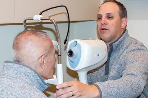 A man with a white device attached to his eye is looking at the man standing in front of him who is holding the device and seems to be explaining something