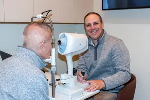 A man is smiling and looking at an eye exam machine while the other man is sitting on a chair in front of him, both of them wearing gray sweaters, and the other man is holding a pen, and they are in an indoor area with a white wall and a mounted TV monitor on it.