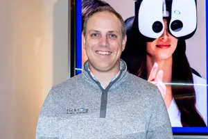 A man wearing a grey sweatshirt is standing in front of a monitor with a smiling woman wearing eye glasses
