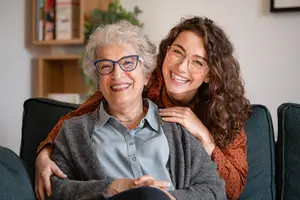 An elderly woman and a young woman are smiling and sitting on a couch with their arms around each other.