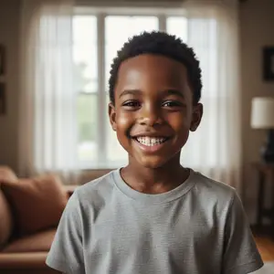 A young boy with a big smile is standing inside a house with a brown couch and white curtain in the background.