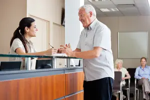 An elderly man and a woman are conversing at the reception desk of an office.