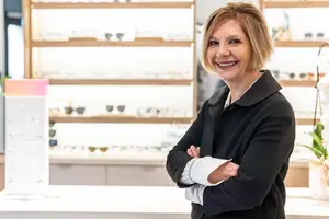 A smiling woman with short blonde hair stands in front of a display of glasses in a shop