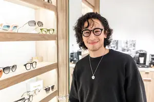 A man wearing glasses is standing in front of a wooden shelf full of sunglasses