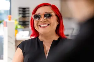 A woman with red hair wearing sunglasses is smiling and looking at a man in a room with a window, potted plants, and shelves in the background.