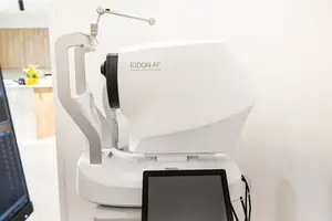 A modern white EIDON AF optometry machine is placed on a white surface, with a monitor screen visible to the left.