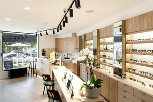 A boutique shop interior featuring a display of glasses and floral arrangements on a white desk, surrounded by wooden cabinets, with a view of a parked car outside through a large window