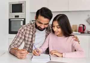 A father helps his daughter with her homework at the kitchen table.
