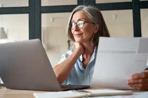 An older woman wearing glasses is sitting on a chair in front of a laptop, looking at a paper and smiling.