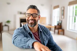 A man wearing glasses and a denim shirt smiles in a living room with a fireplace and windows in the background.