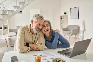 A couple sitting at a table with a laptop and a bowl of food, smiling at each other.