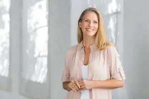 A woman with blonde hair is smiling and standing in front of a white wall with windows